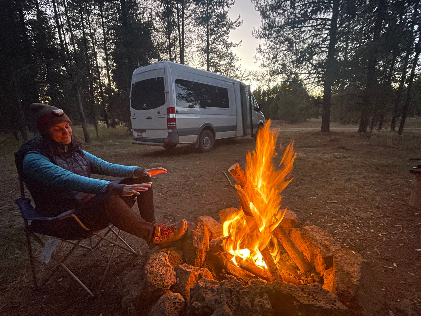 RV rental adventure: woman warms hands by a crackling campfire, her camper van parked in a forest at dusk.