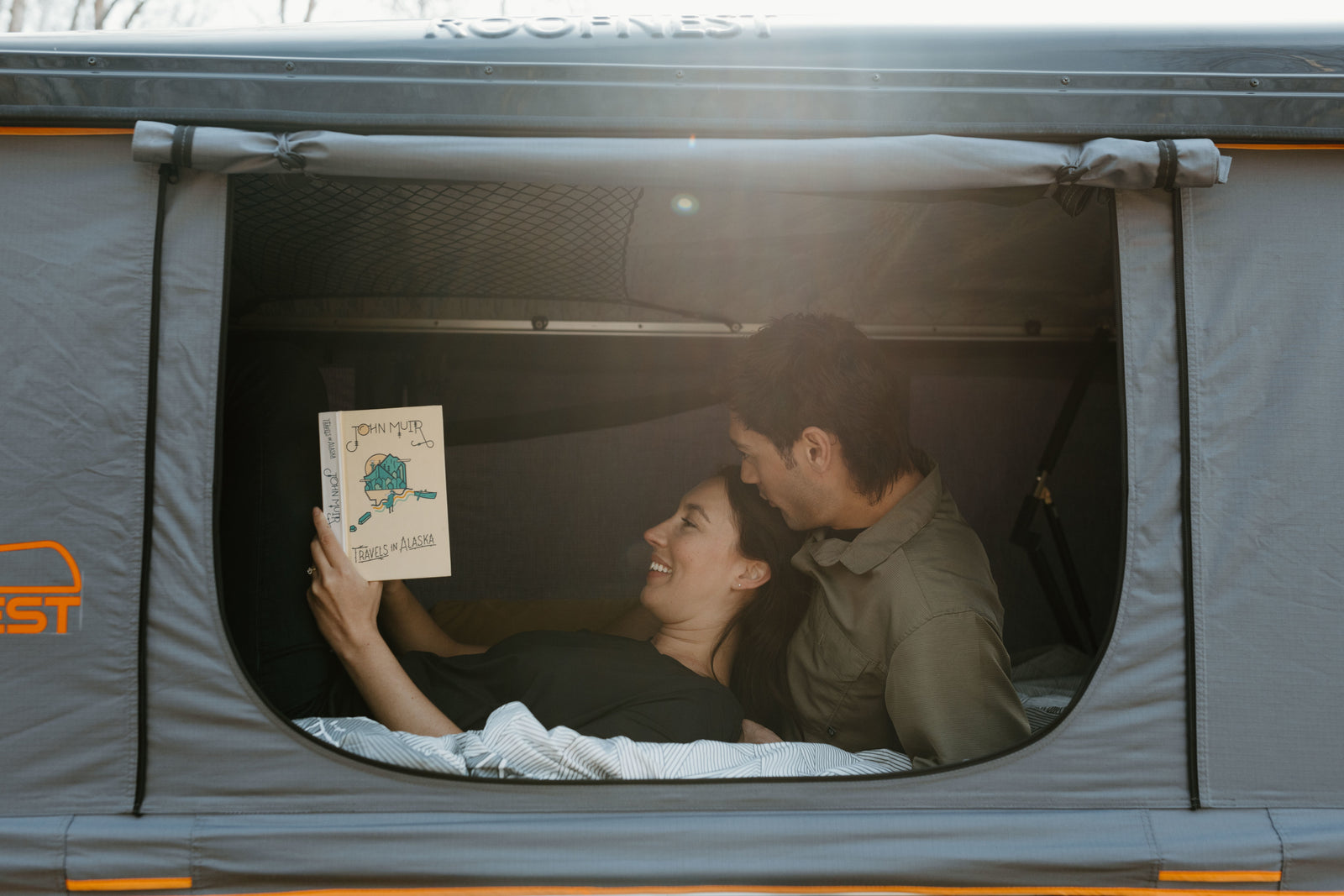 A happy couple relaxes in a Roofnest rooftop tent, reading "Travels in Alaska," ideal for any RV rental adventure.