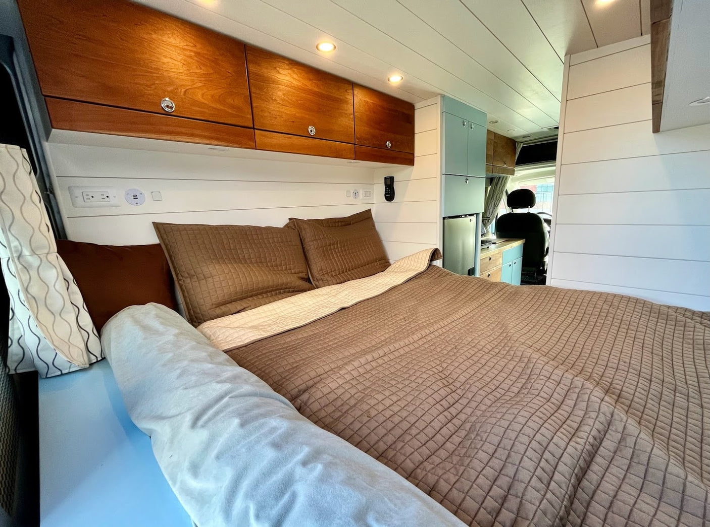 Cozy RV rental bedroom with warm wooden cabinets, white shiplap, and a comfy bed. A light blue kitchenette is visible. Ideal for travel.