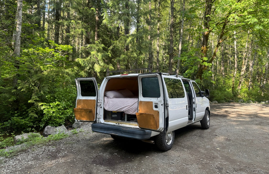 Cozy white RV rental camper van parked in a vibrant green forest, doors open revealing a bed. Perfect for a road trip!
