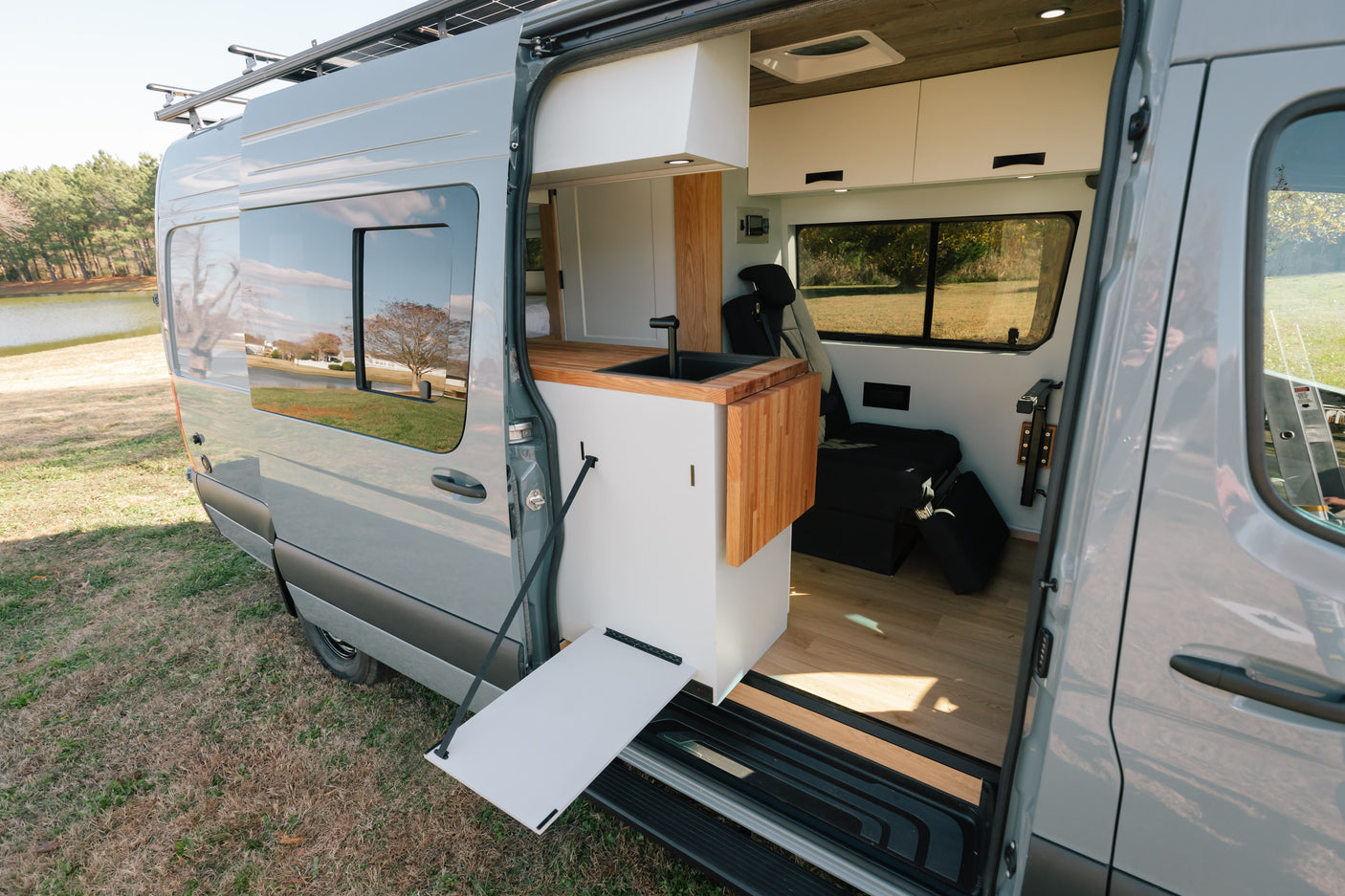 Stylish RV rental: Grey camper van interior with kitchen, flip-out table, and living area, parked by a scenic lake.