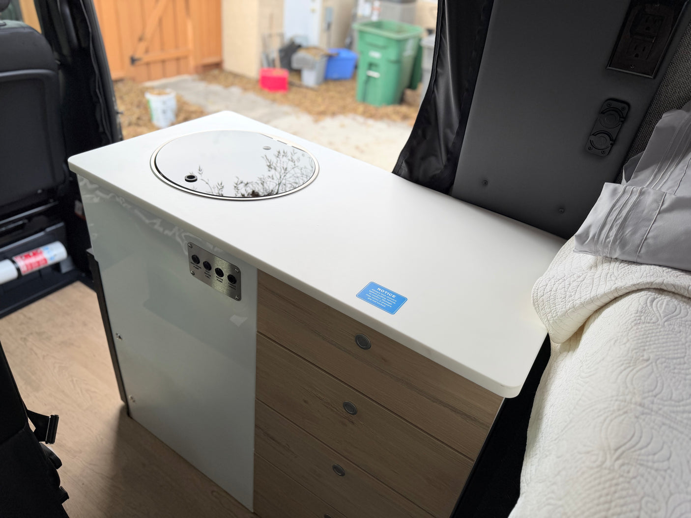 RV rental camper van interior featuring a kitchenette with sink, control panel, wood drawers, and a bed. Outside is visible.