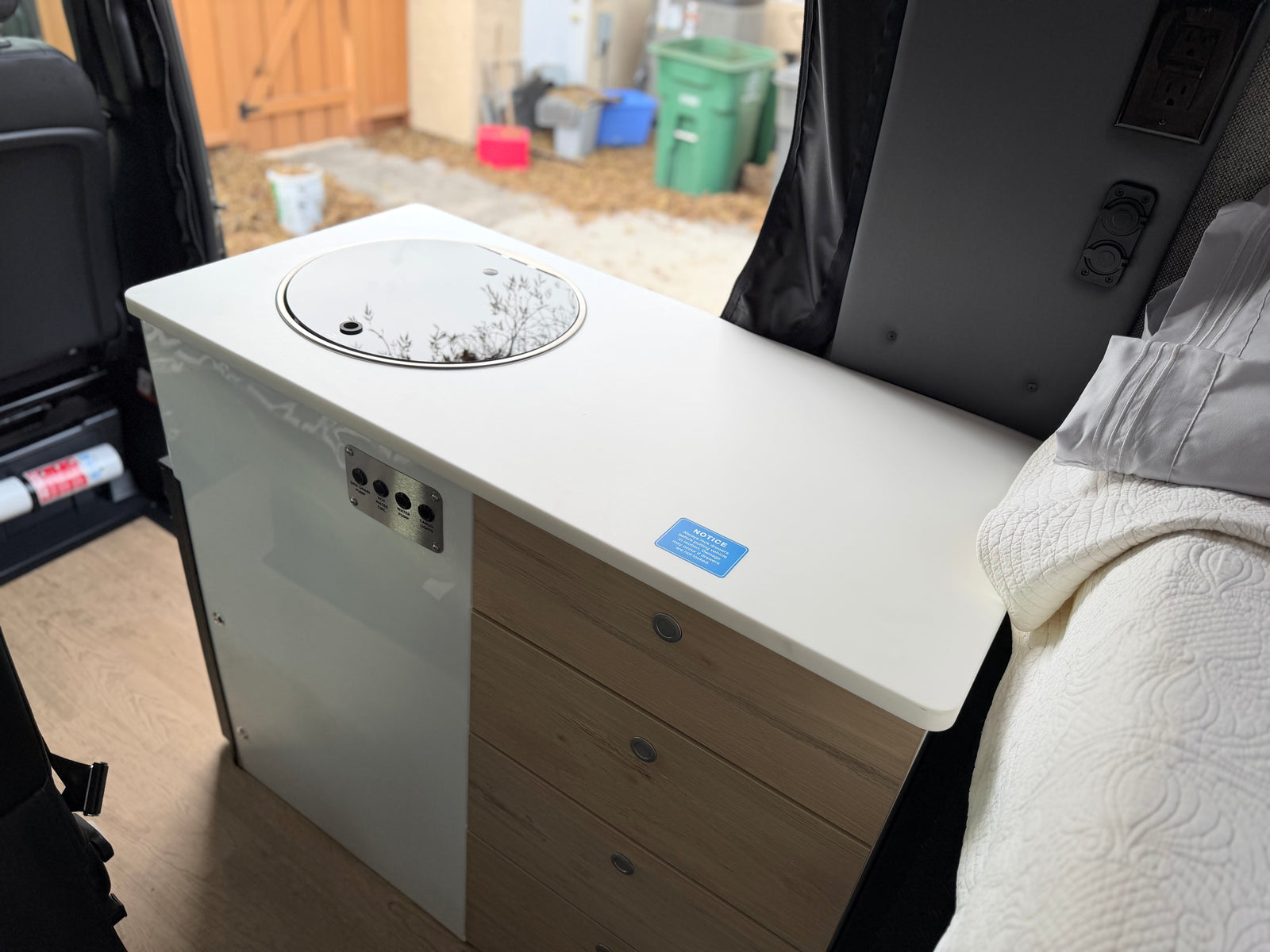 RV rental camper van interior featuring a kitchenette with sink, control panel, wood drawers, and a bed. Outside is visible.
