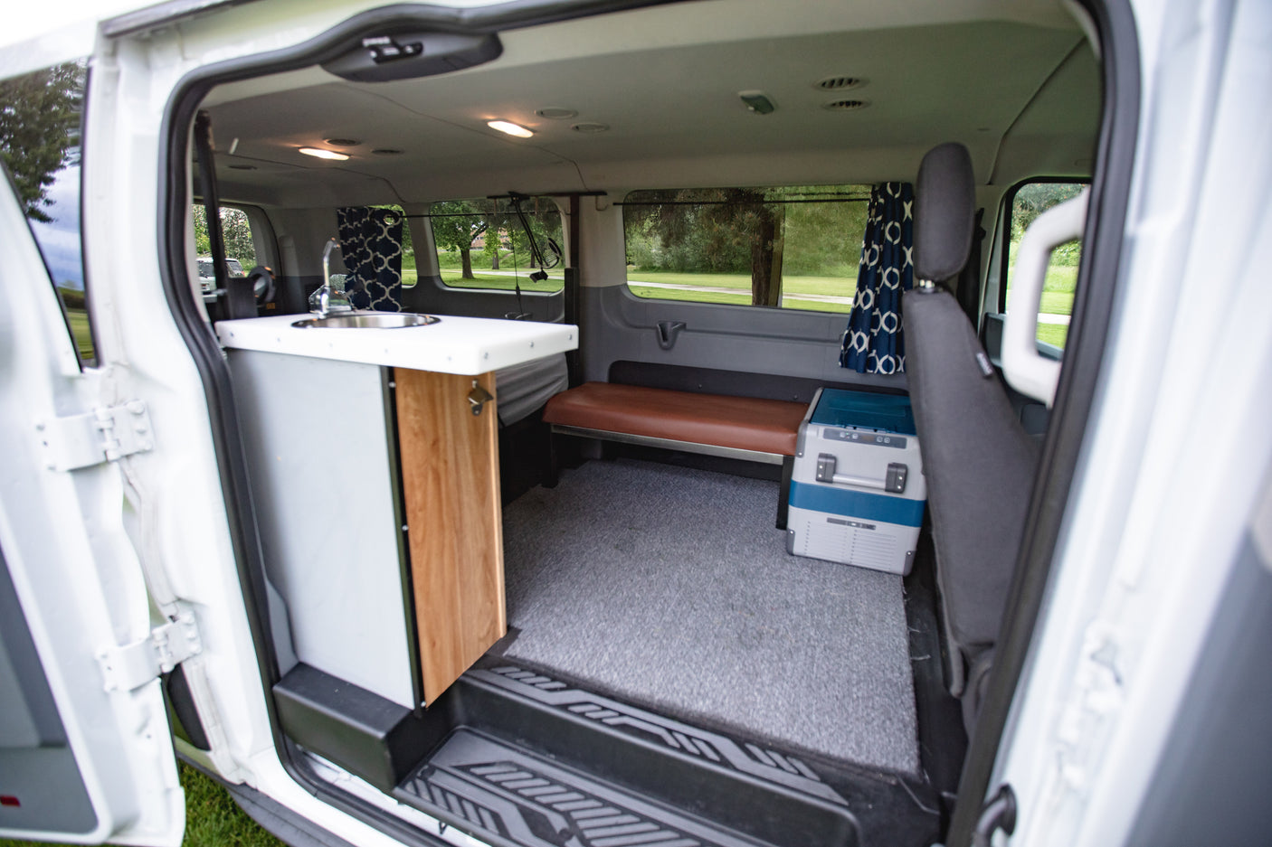 Inside this RV rental camper van: a compact kitchen with sink, bench seating, and portable cooler on grey carpet.