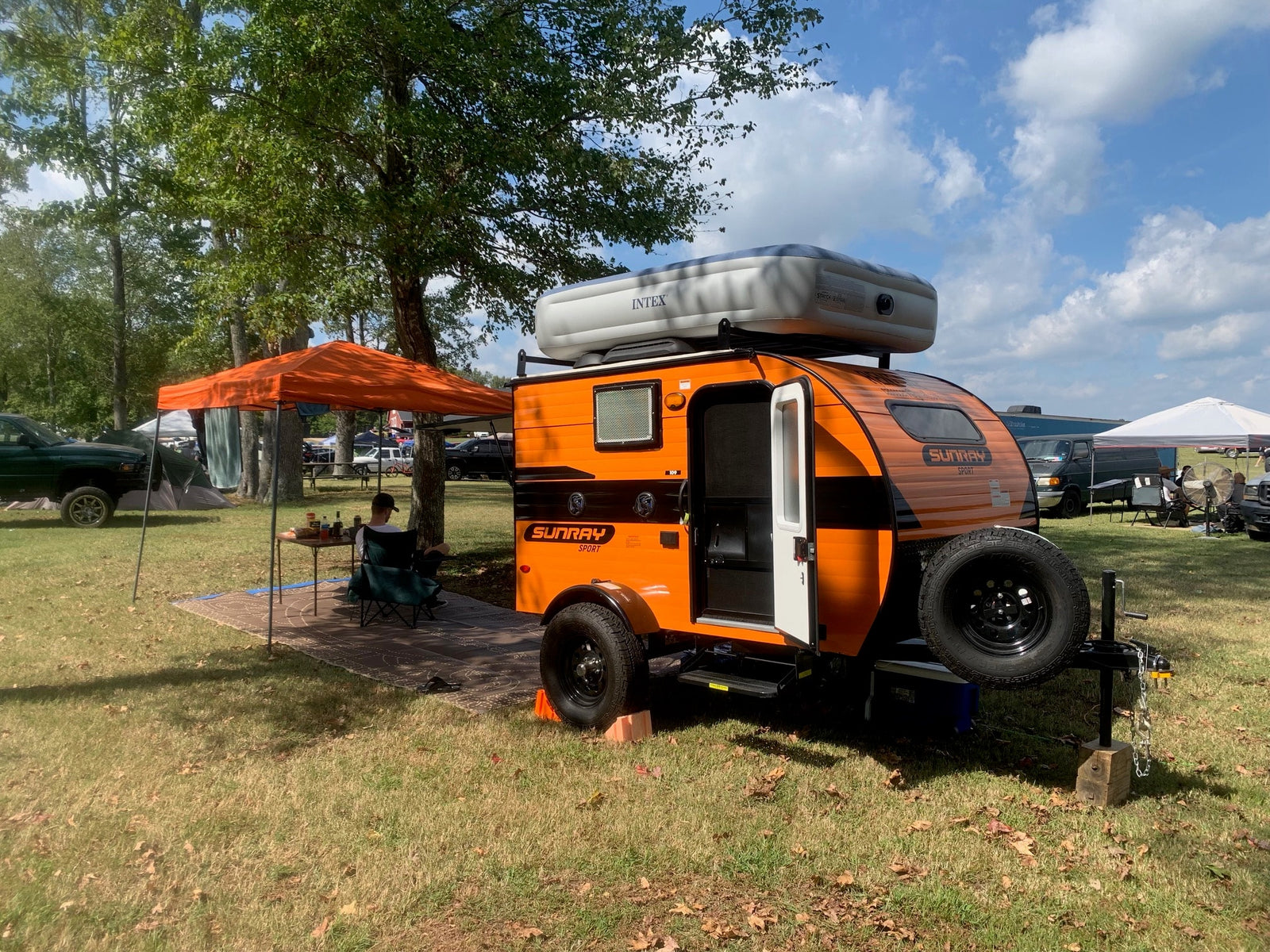 Vibrant orange Sunray teardrop RV rental with air mattress on top, alongside a canopy at an outdoor event.