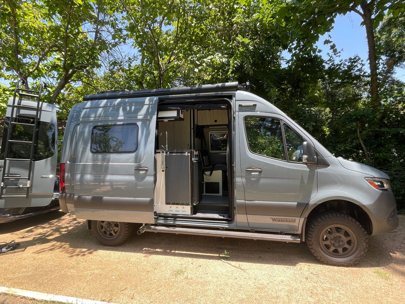 Explore in a Winnebago camper van RV rental. Side door open, revealing a functional interior amidst green trees.