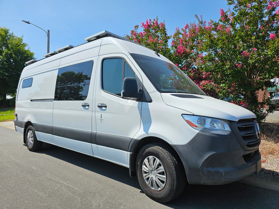 White Mercedes Sprinter RV rental van with solar panels, parked by pink-flowering trees on a sunny day.