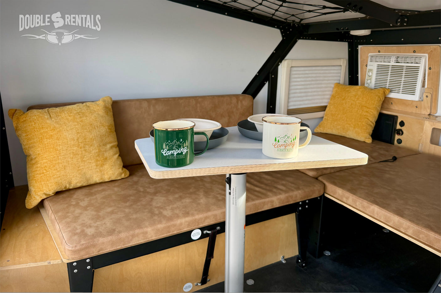 Inside this modern RV rental, find a comfortable dining nook with tan benches, a table, enamel mugs, and a window AC unit.