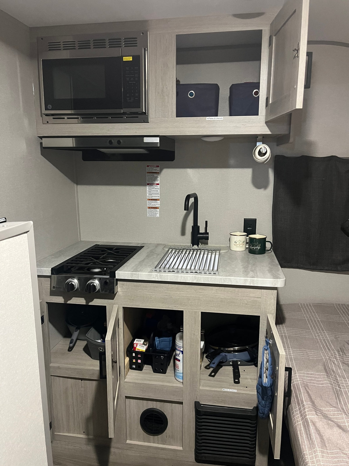 RV rental kitchen view: GE microwave, two-burner stove, sink with drying rack, and accessible cabinet storage. — photo 6