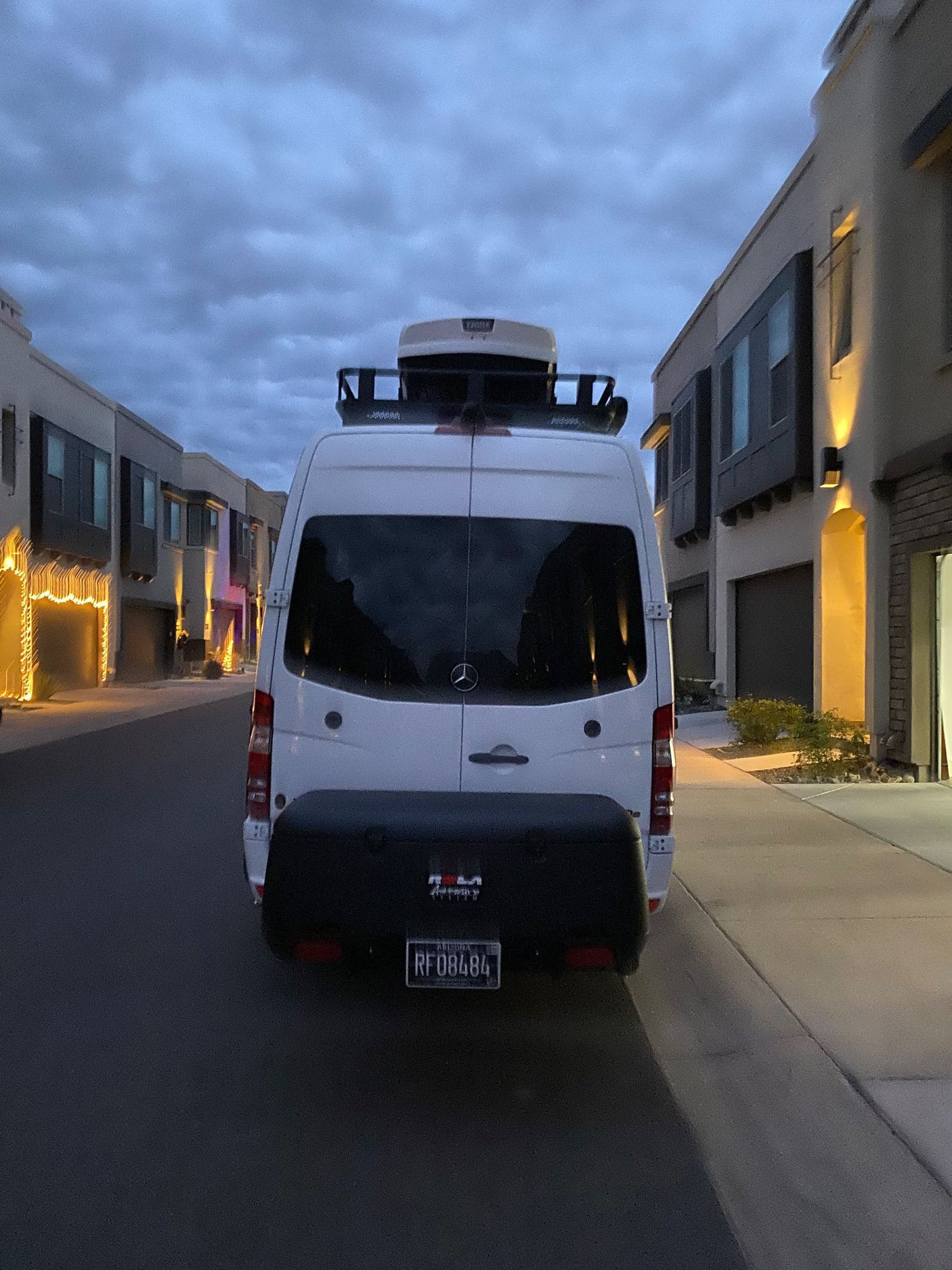 White Mercedes Sprinter RV rental van with cargo box & rooftop carrier, parked in a modern neighborhood at dusk.