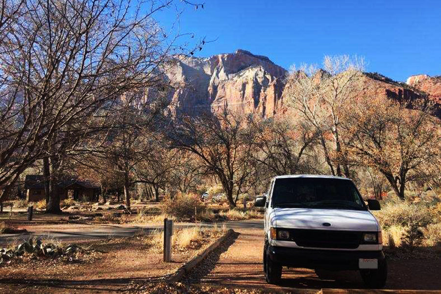 White RV rental van at a scenic park campsite, with majestic red rock mountains and leafless trees under blue sky.