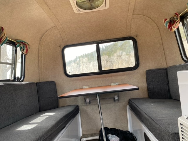 Inside an RV rental: a compact dinette with two grey benches, a central table, and scenic windows overlooking trees.