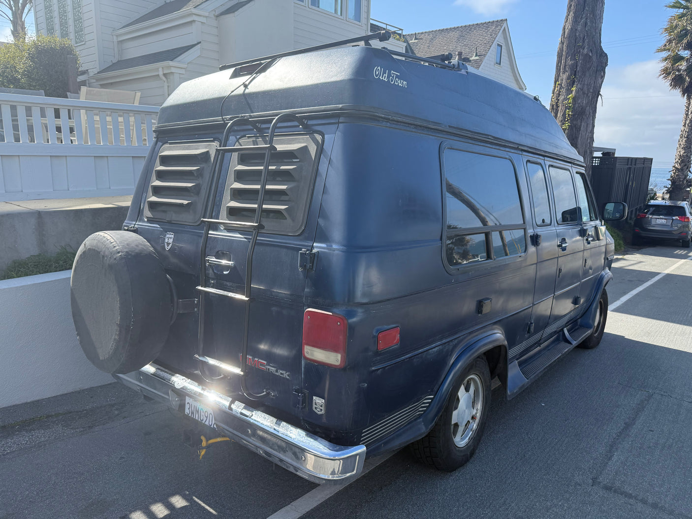 Dark blue "Old Town" GMC conversion van, an RV rental with a ladder & spare tire, parked by the ocean.