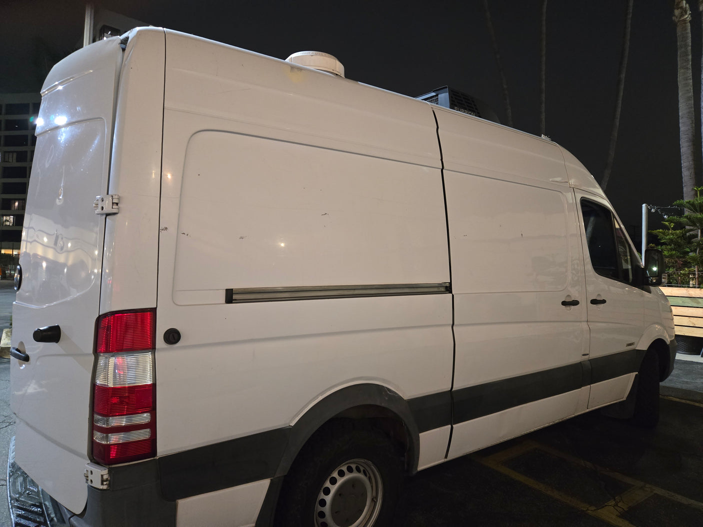 White RV rental van, converted into a camper with roof AC and vent, parked at night, rear-side view.