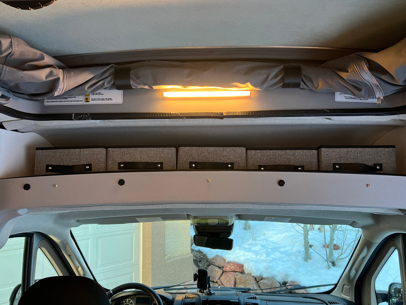 Pop-top RV rental interior: overhead light, storage bins, and roof warnings above the dashboard, showing a snowy view.