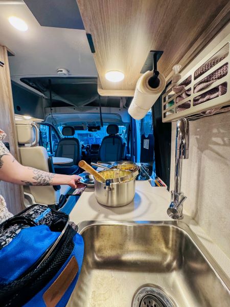 Inside a cozy RV rental: a person prepares food in the compact kitchen featuring a sink, pot, and overhead storage.