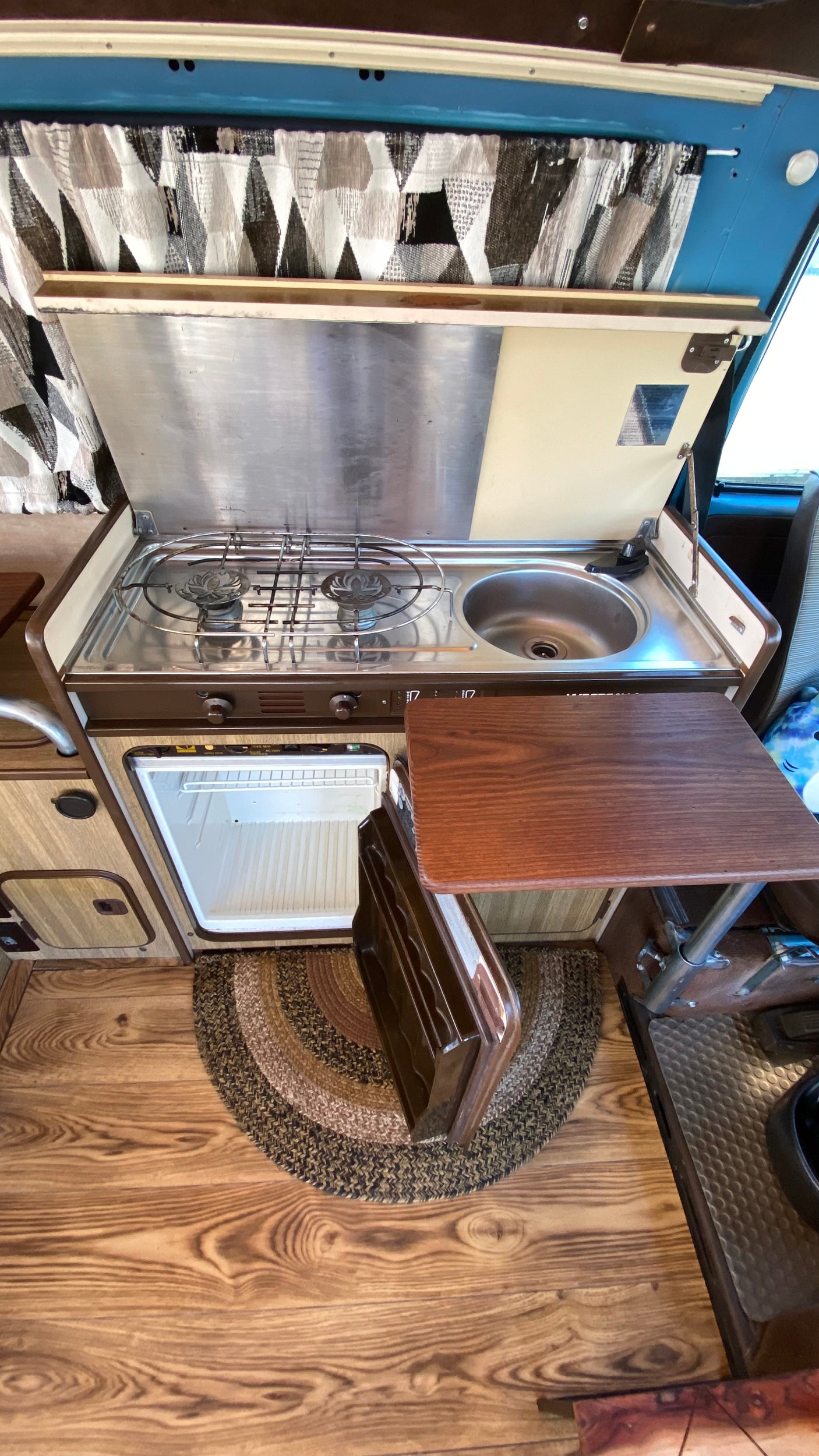 Detailed view of an RV rental's compact kitchen: stainless steel stove, sink, open mini-fridge, and wooden table.