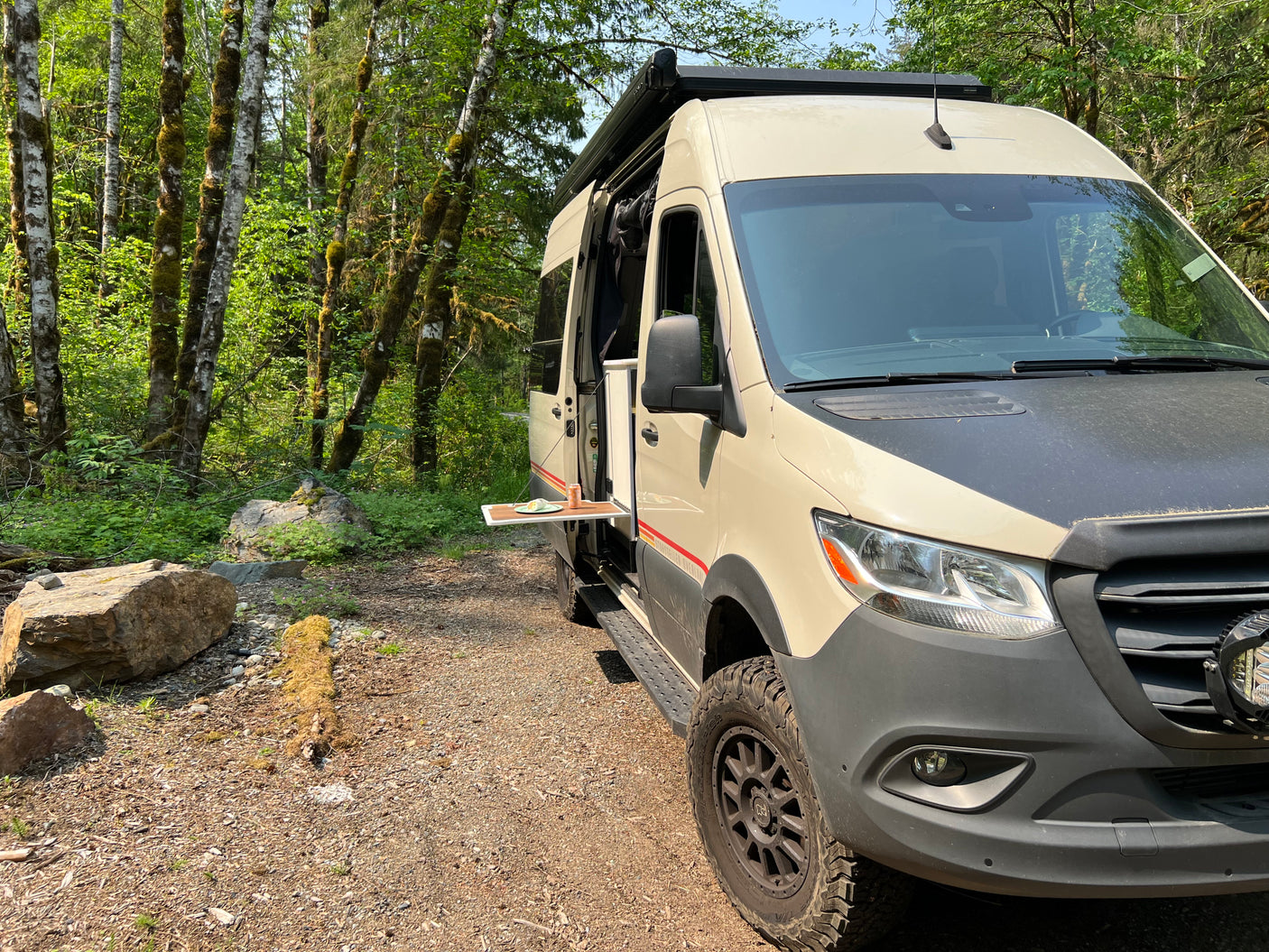 Explore nature with an RV rental! This camper van is parked in a lush forest with a side table extended for a meal.