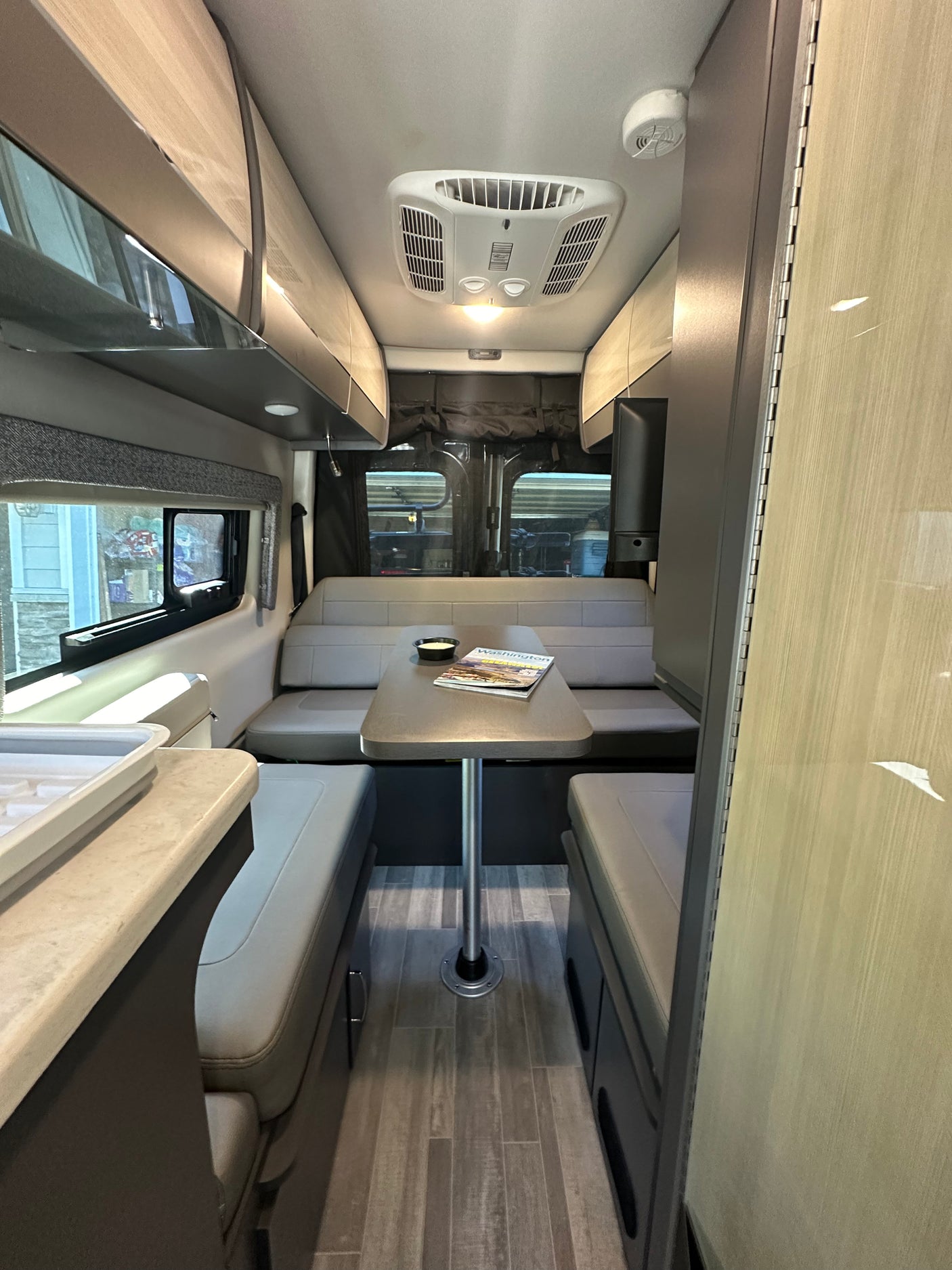 RV rental interior featuring a central dining table with benches, glimpsed kitchen, and overhead cabinets for compact living.