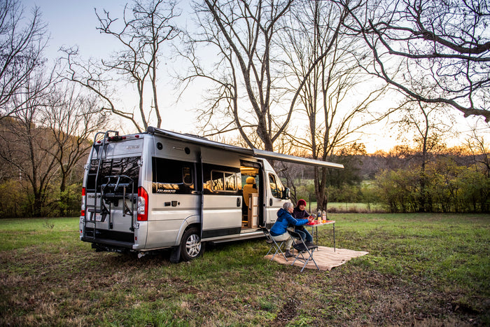 An RV rental camper van parked in a grassy field at sunset, with two people enjoying dinner outside under an awning.