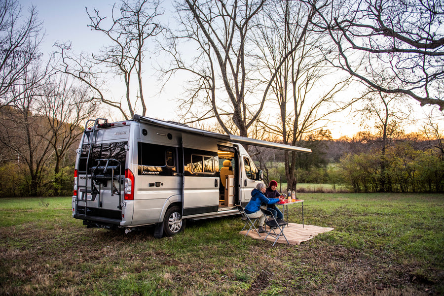 An RV rental camper van parked in a grassy field at sunset, with two people enjoying dinner outside under an awning.