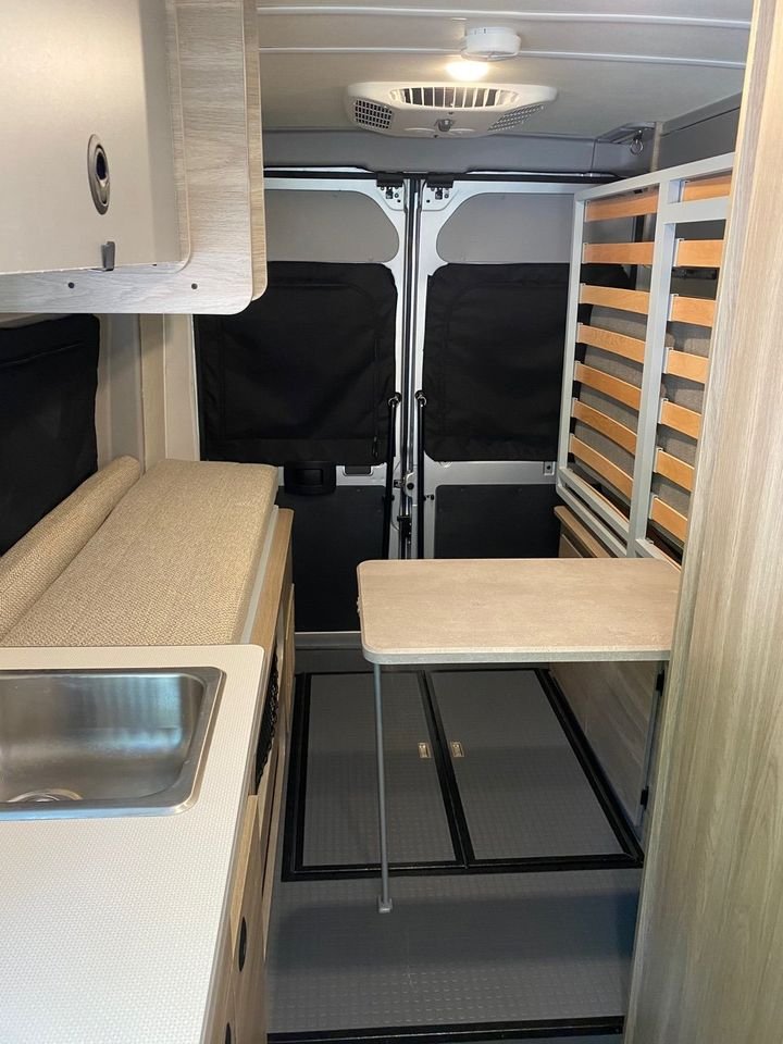 Spacious RV rental interior: kitchenette with sink, sofa-bed, compact table, and slatted bed frame near rear doors.