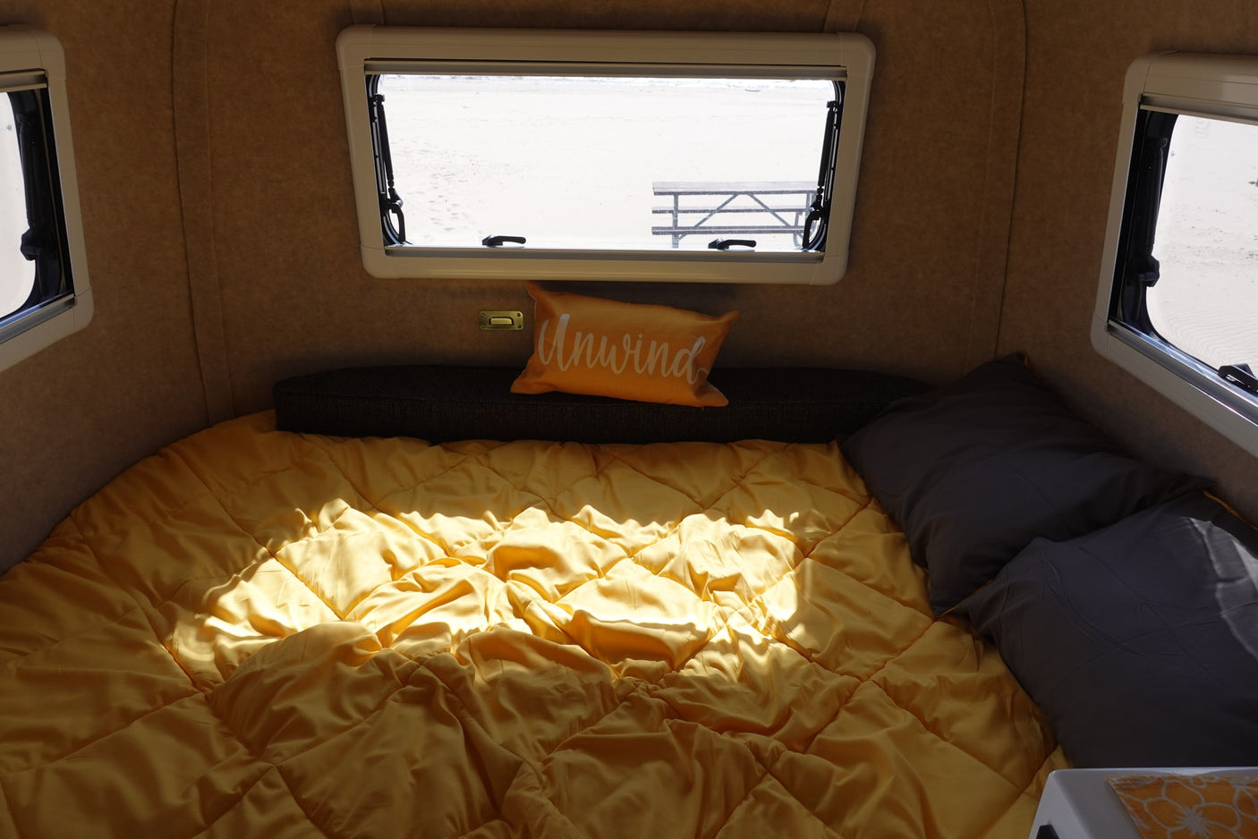 Cozy RV rental interior with a sunny bed, "Unwind" pillow, and a view of a beach and picnic table through the window.