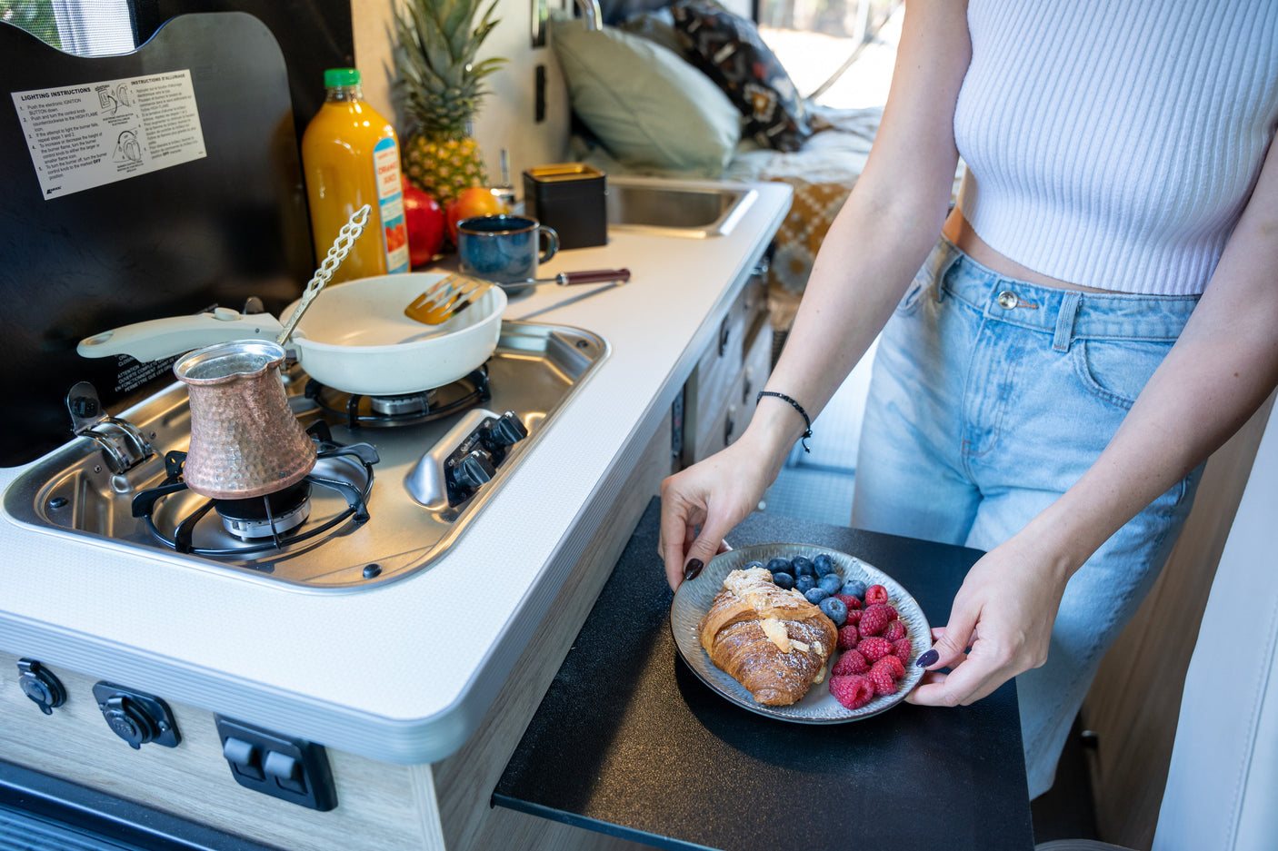 A woman prepares breakfast in a compact RV rental kitchen, with coffee brewing and fresh fruit on a plate. Ideal for travel.