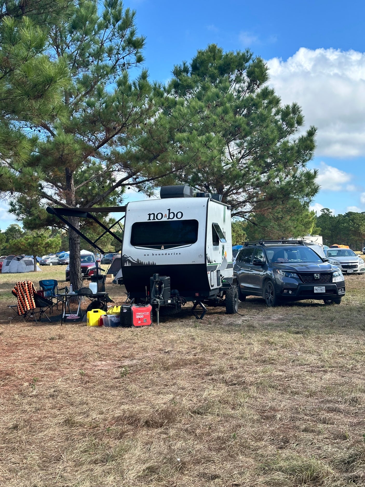 Scenic RV rental campsite featuring a white 'no bo' travel trailer, SUV, and outdoor gear under a large pine tree.