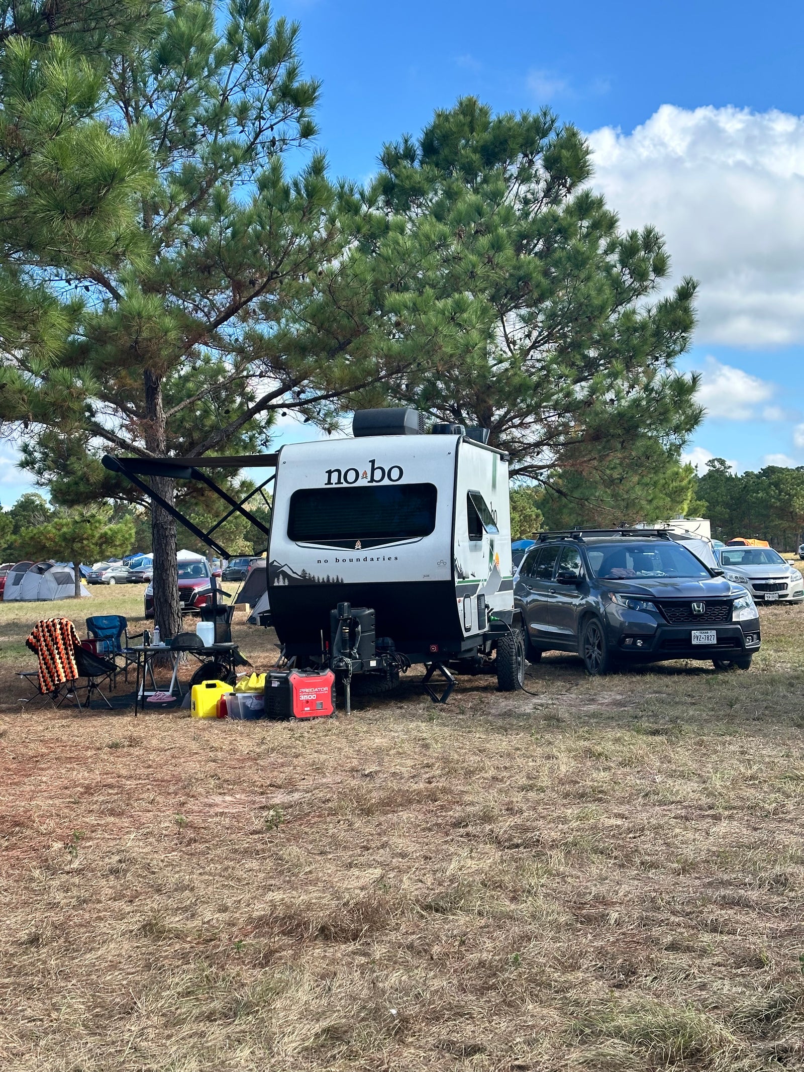 Scenic RV rental campsite featuring a white 'no bo' travel trailer, SUV, and outdoor gear under a large pine tree.
