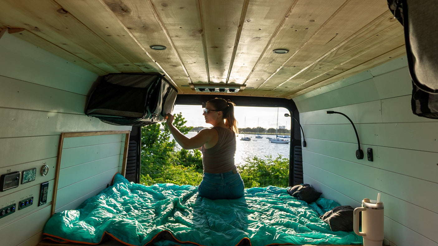 Inside an RV rental, a woman on a teal-covered bed reaches overhead storage, with a scenic lake and boats outside.