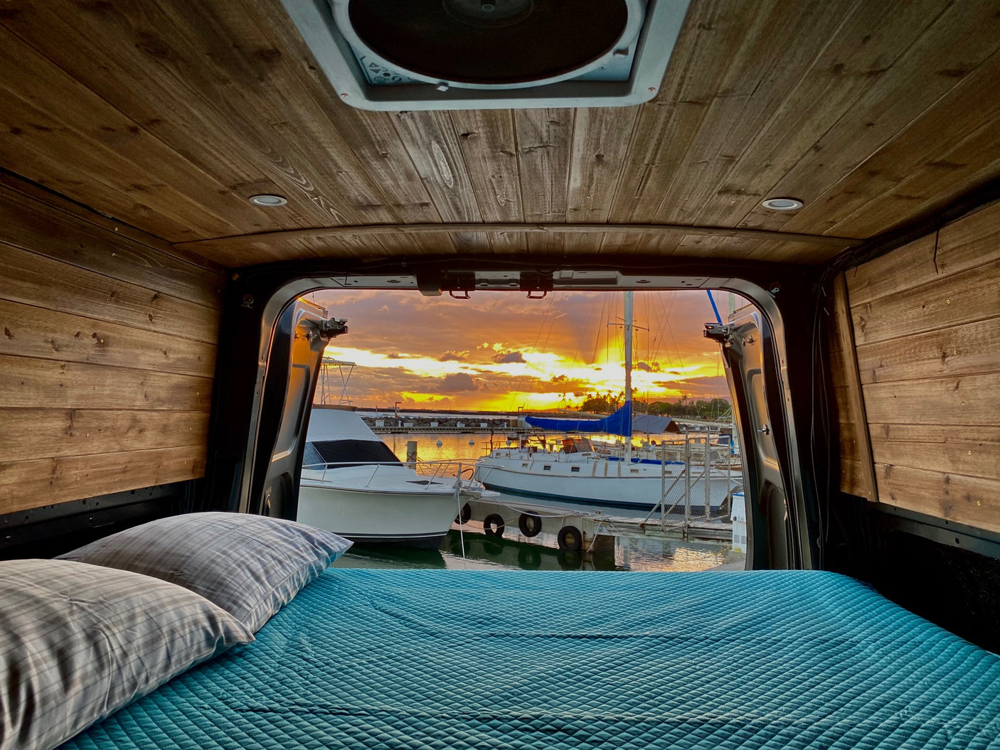 Cozy rv rental bed looking out to a stunning orange sunset over a marina filled with boats. Perfect for van life adventures.