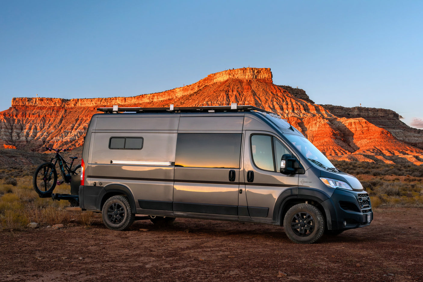 Gray RV rental van with mountain bike on a dirt road, facing sunlit red mountains under a blue sky. Perfect for adventure.