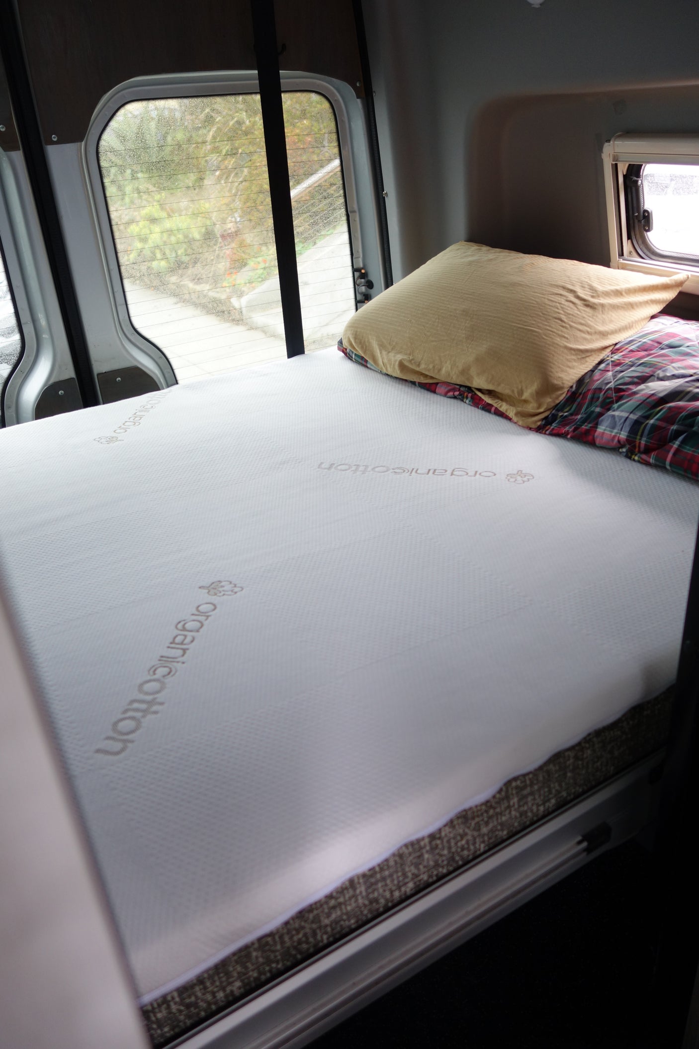 RV rental's cozy bed: a white organiCotton mattress, pillow, and plaid blanket inside a camper van, with window views.