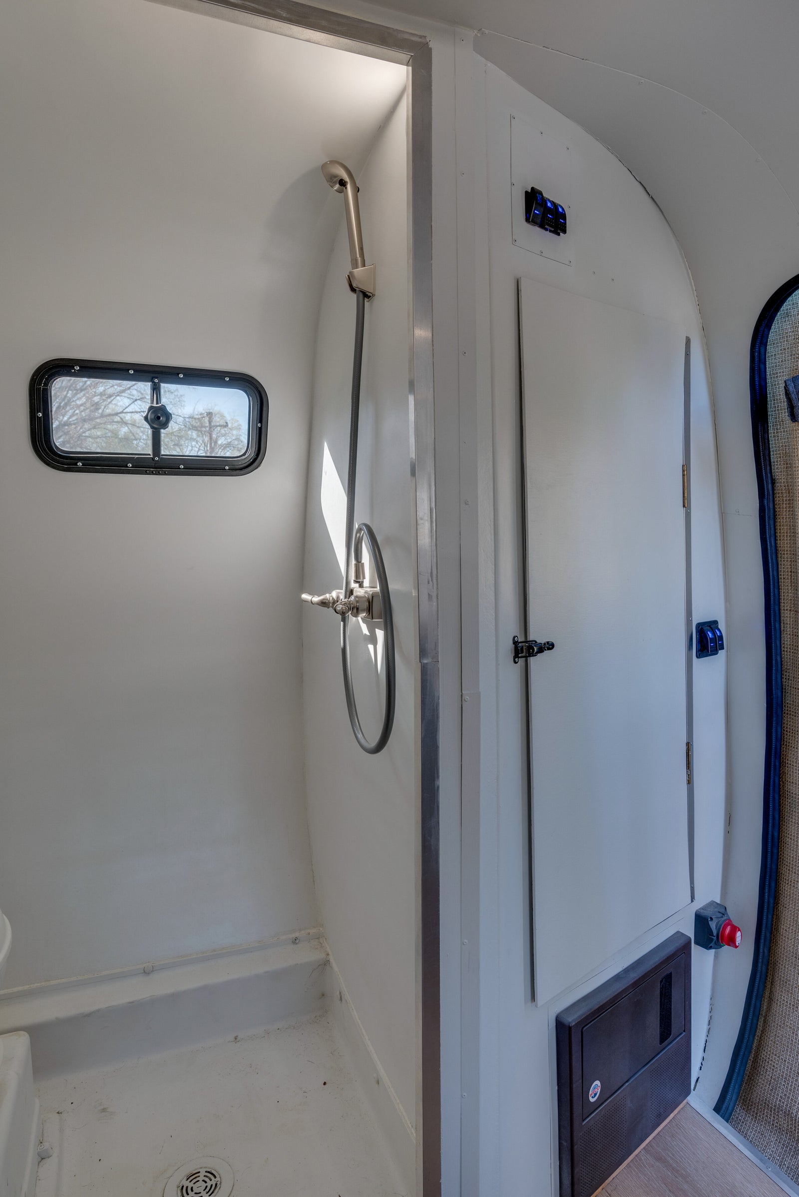 Compact rv rental interior: bright white shower with window and controls, adjacent panel with blue switches, and electrical access.