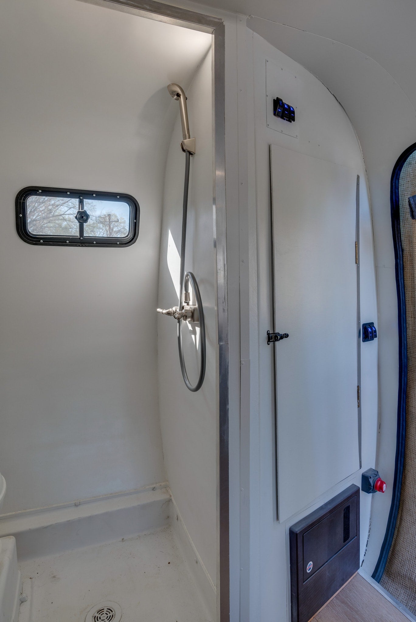 Compact rv rental interior: bright white shower with window and controls, adjacent panel with blue switches, and electrical access.