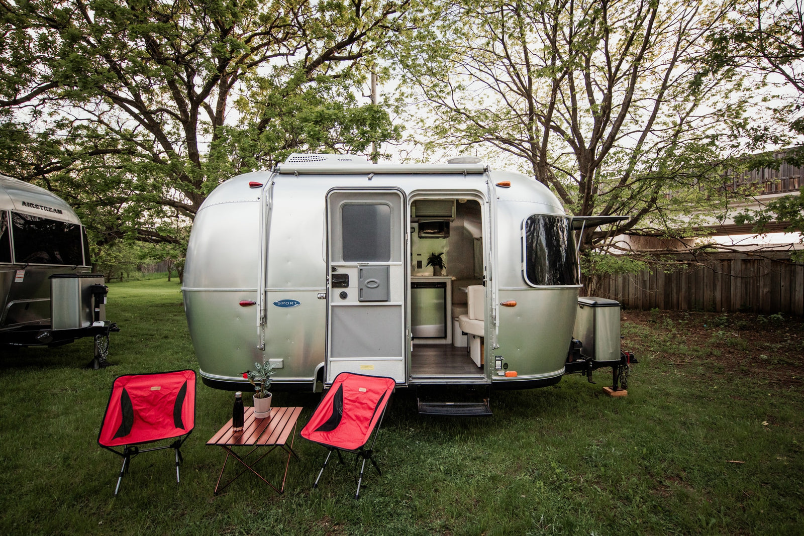 Silver Airstream rv rental with open door showing its interior, plus red chairs & a table outside in a grassy, treed setting.