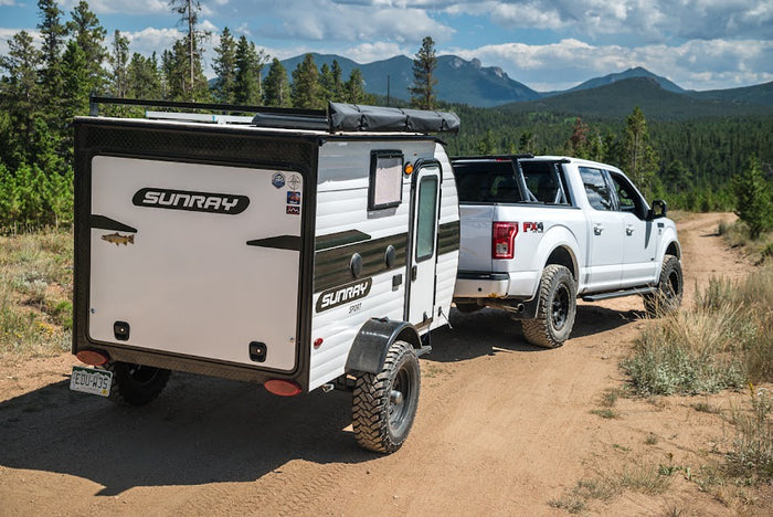 perfect for camping adventures in scenic nature.|A white pickup truck tows a Sunray teardrop RV down a dirt mountain trail. Perfect for your next RV rental adventure.|Explore the outdoors with this off-road rv rental: a white Sunray Sport travel trailer towed by a Ford F-150 truck.|White truck towing RV rental on a dirt road through a vast forest. Mountains in distance under a cloudy sky.|Enjoy an RV rental: Sunray Sport trailer parked with an open door