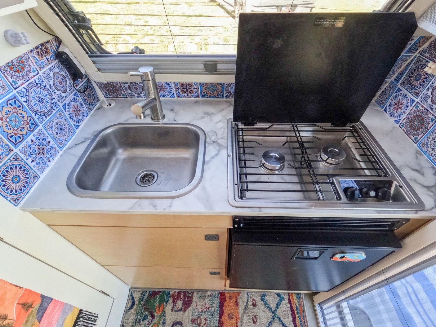 Compact RV rental kitchen: stainless sink, Dometic stove, fridge, and vibrant patterned tile backsplash under a window.