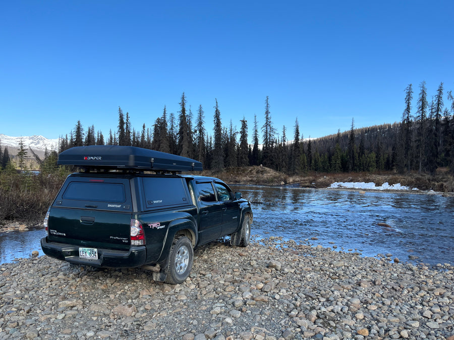 Overland RV rental: A pickup truck with a rooftop tent by a river, forests, and snowy mountains. Ready for Alaska adventure & camping.