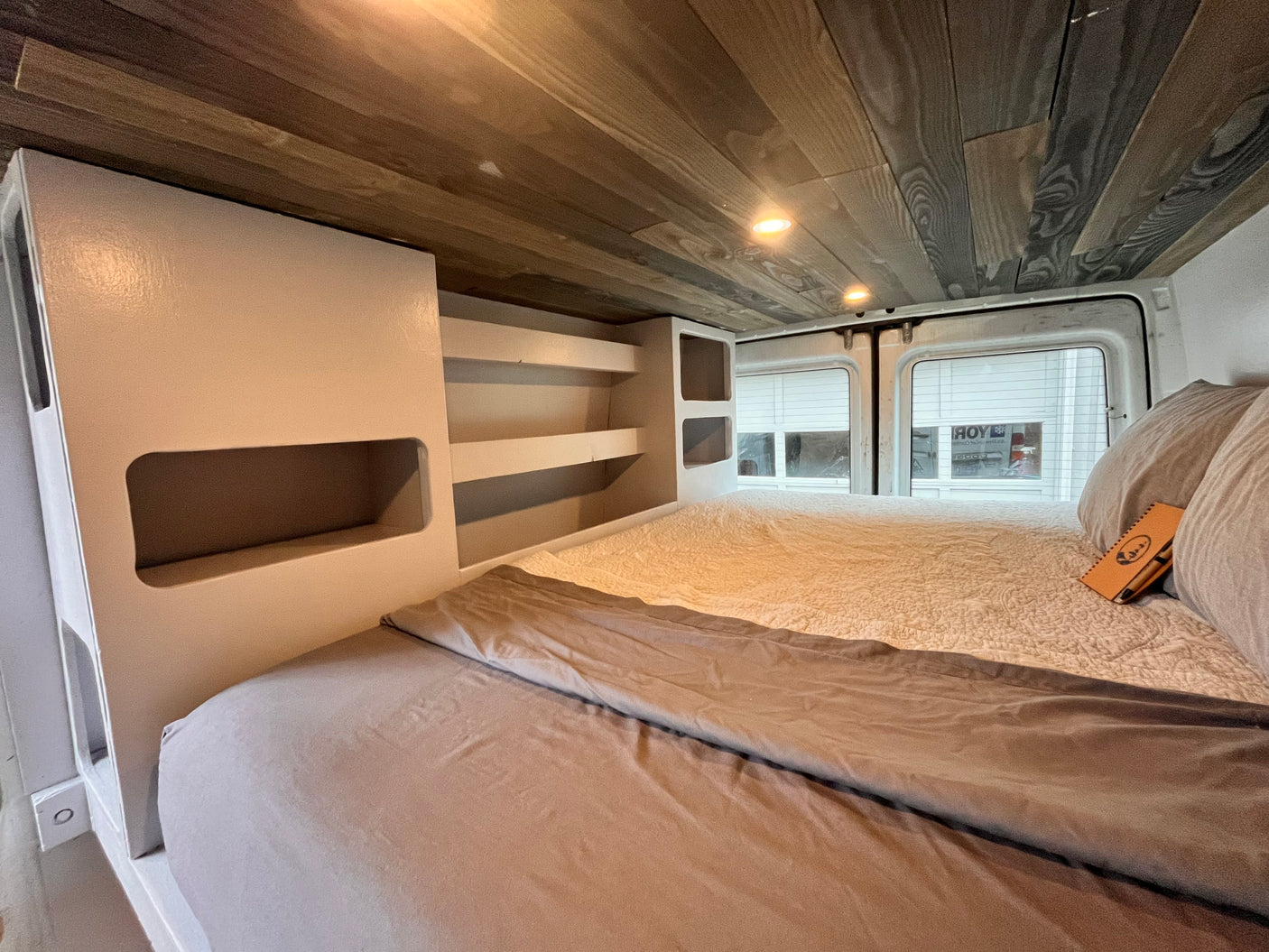 RV rental: Inside a comfortable camper van bedroom with a bed, light bedding, wood-paneled ceiling, and shelving.