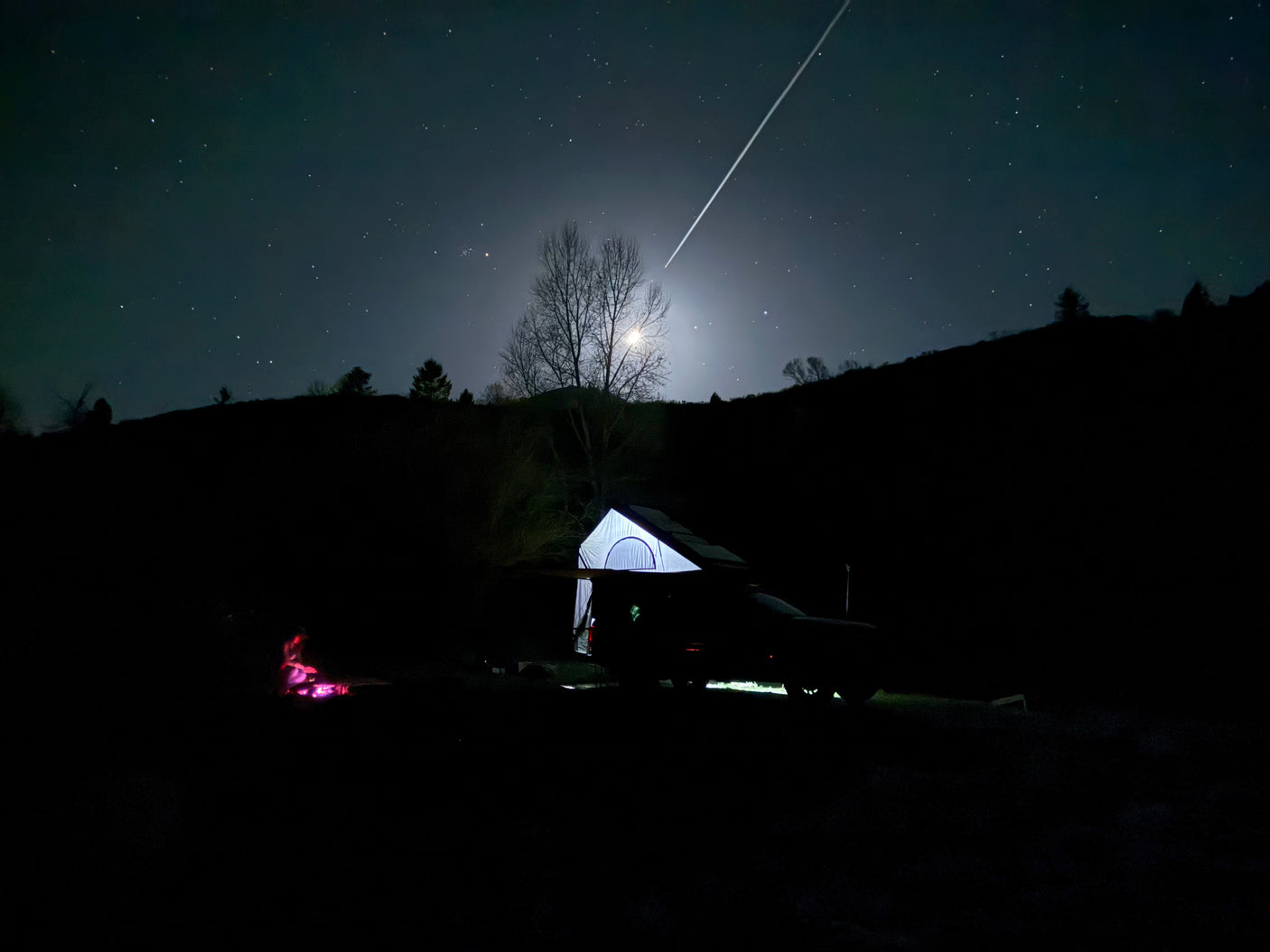Experience starry nights: a moonlit camping vehicle with tent (like an RV rental) under a plane trail & glowing campfire.
