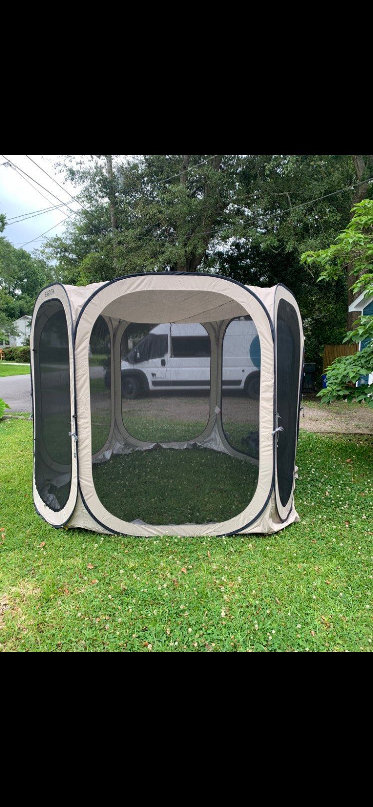 Tan pop-up screen house on grass with screened walls, set up outdoors. A white RV rental van is parked in the background.