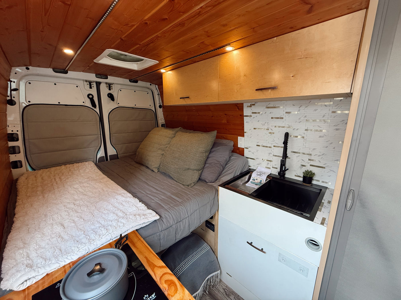 Cozy RV rental interior featuring a comfortable bed, modern kitchenette with sink and backsplash, wood-paneled ceiling, and storage.