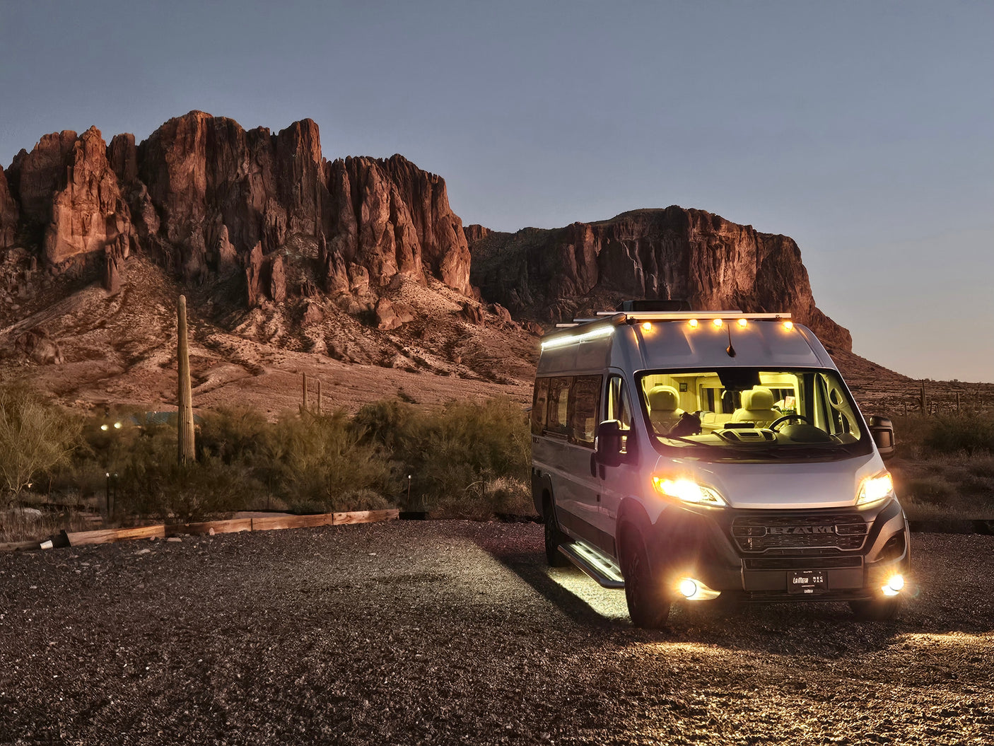 An RV rental camper van, lit up at dusk, parked on a gravel lot with majestic desert mountains in the background.