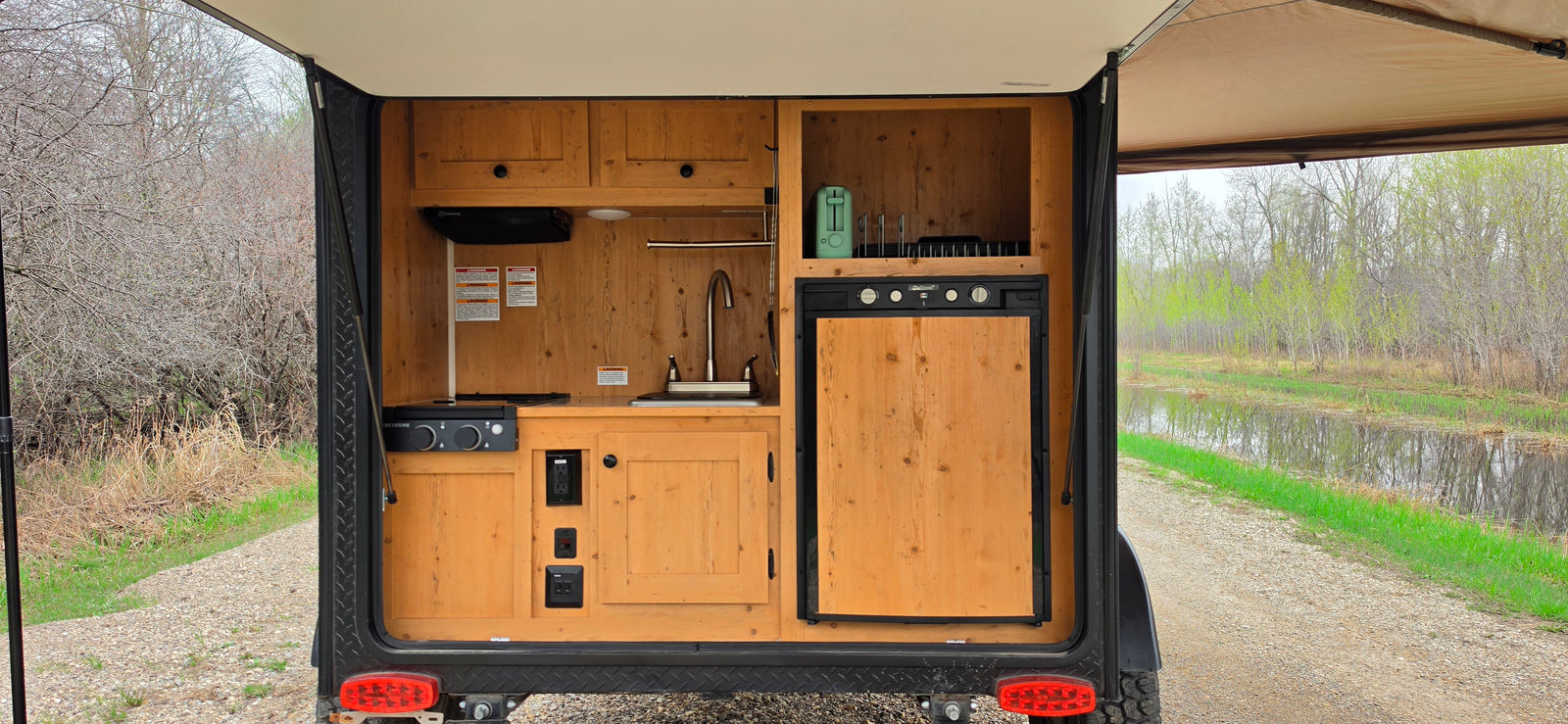 This RV rental teardrop camper boasts an exterior kitchen with a fridge, sink, and stove, ideal for roadside cooking near water.