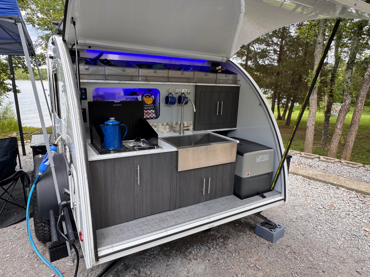 Compact RV rental teardrop camper's illuminated outdoor kitchen with stove, sink, & fridge, set up by a lake.