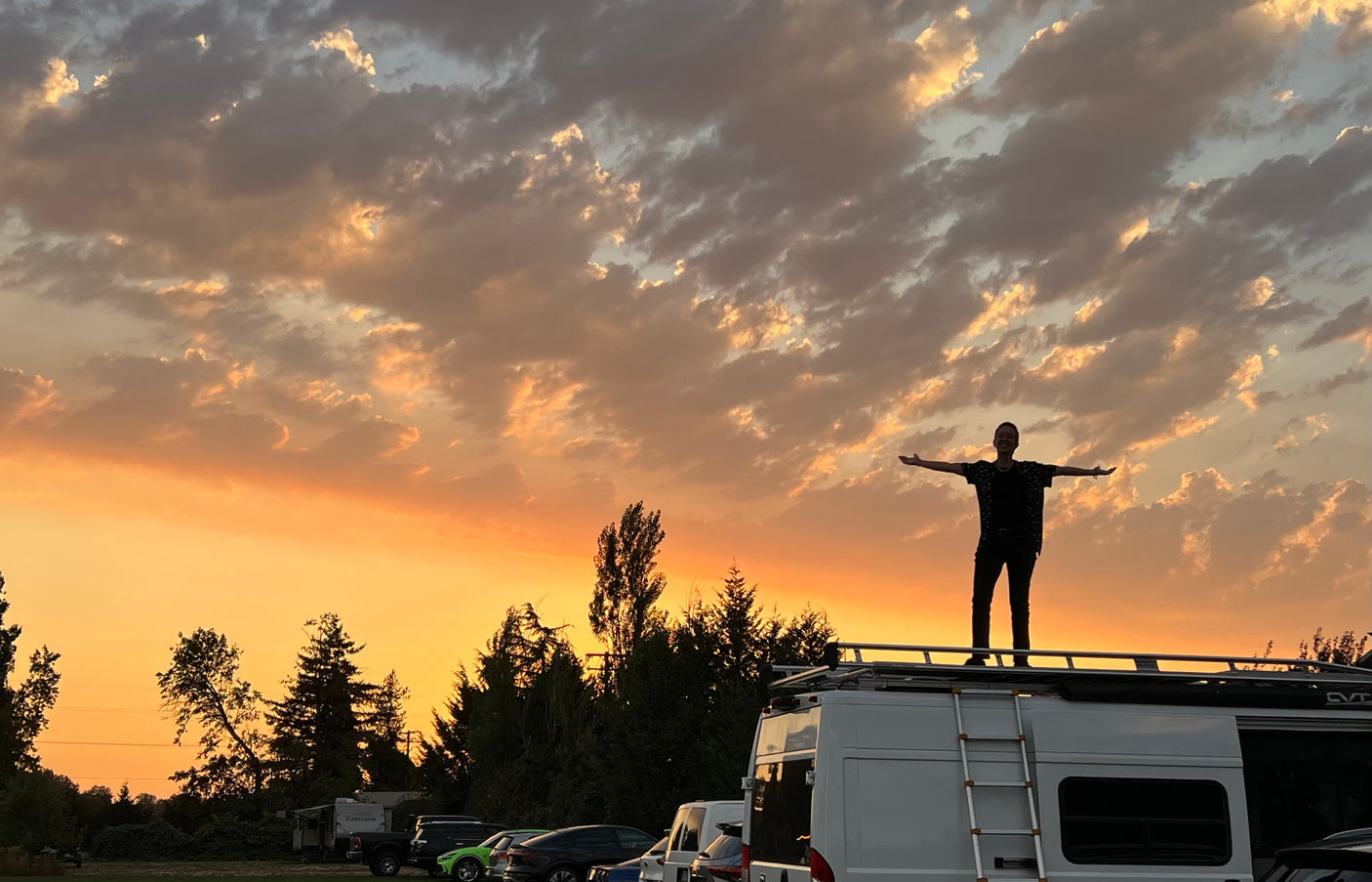 Experiencing golden hour from an RV rental! Person stands silhouetted on the roof, arms outstretched, admiring the colorful sky.
