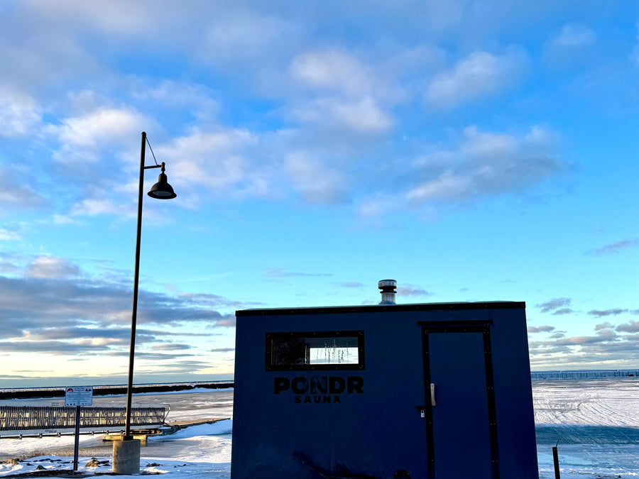 Dark blue Pondr sauna, a unique outdoor structure like an rv rental, on an icy lake. Winter scene with a lamp post and blue sky.