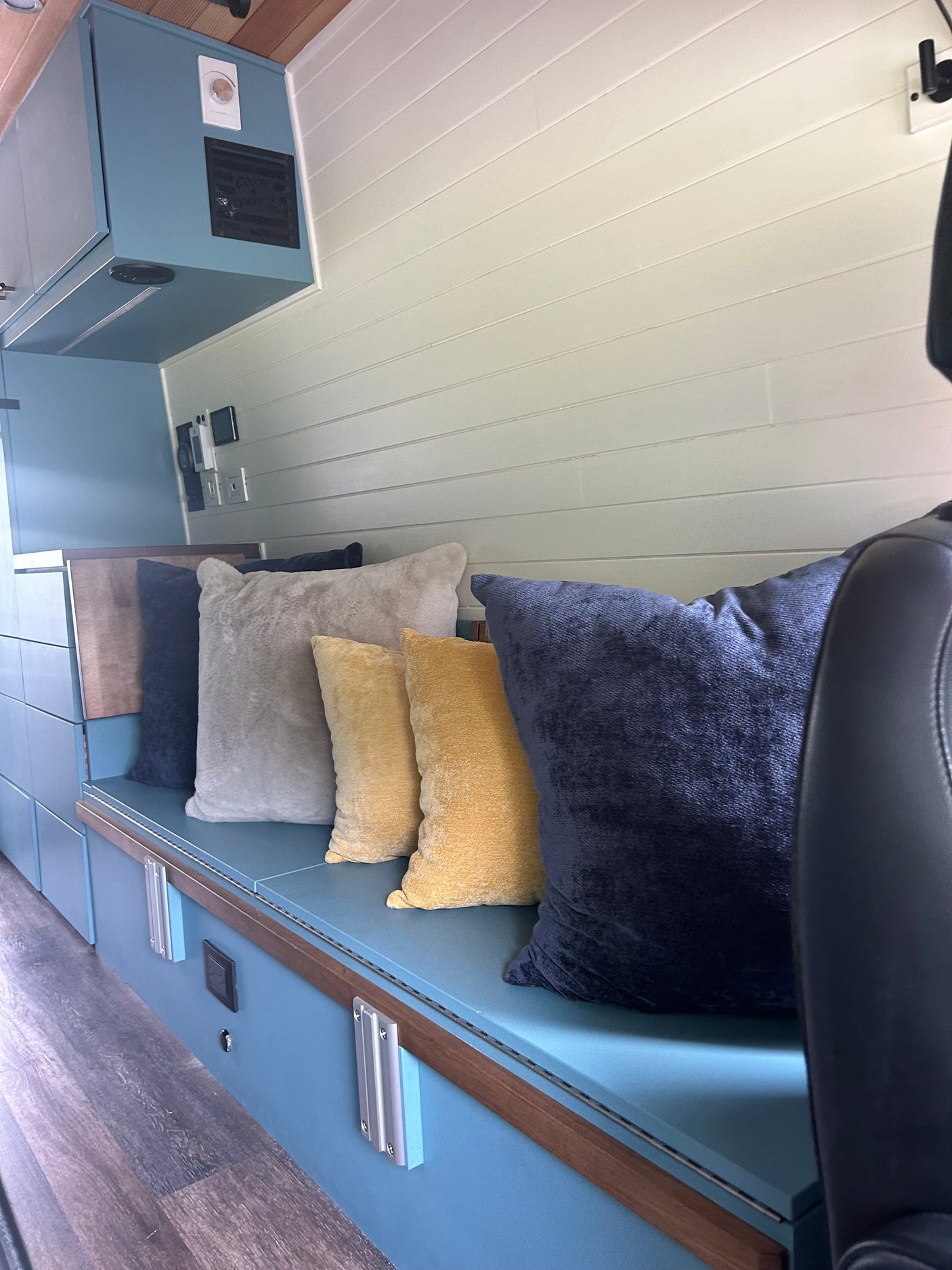 Cozy RV rental interior featuring a blue bench seat with assorted pillows, overhead cabinets, and wood-look flooring for comfortable travel.